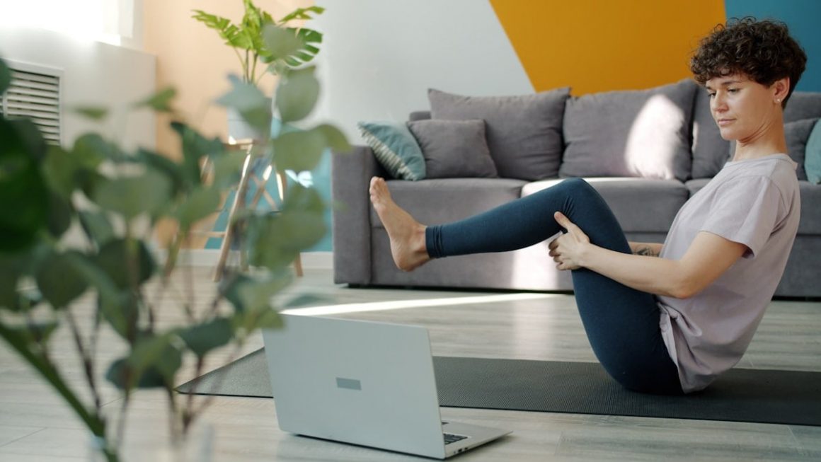 Flexible girl is learning yoga online using laptop watching tutorial in internet doing asanas sitting on mat in flat. Modern technology, youth and relaxation concept.