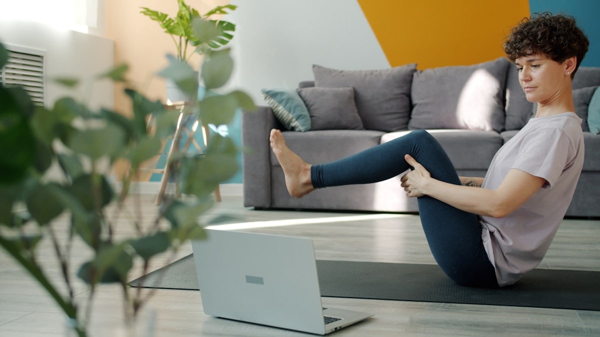 Flexible girl is learning yoga online using laptop watching tutorial in internet doing asanas sitting on mat in flat. Modern technology, youth and relaxation concept.